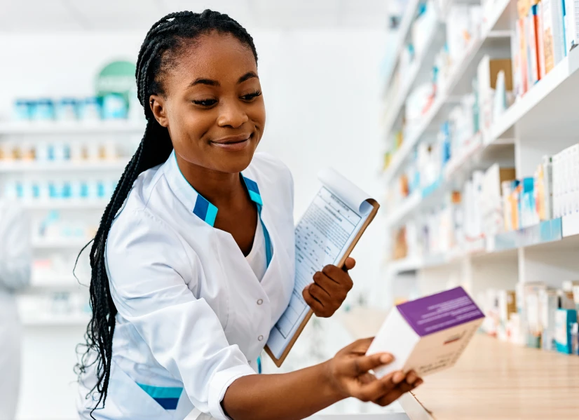 A pharmacist in a lab coat with a clipboard looking at a box of medication
