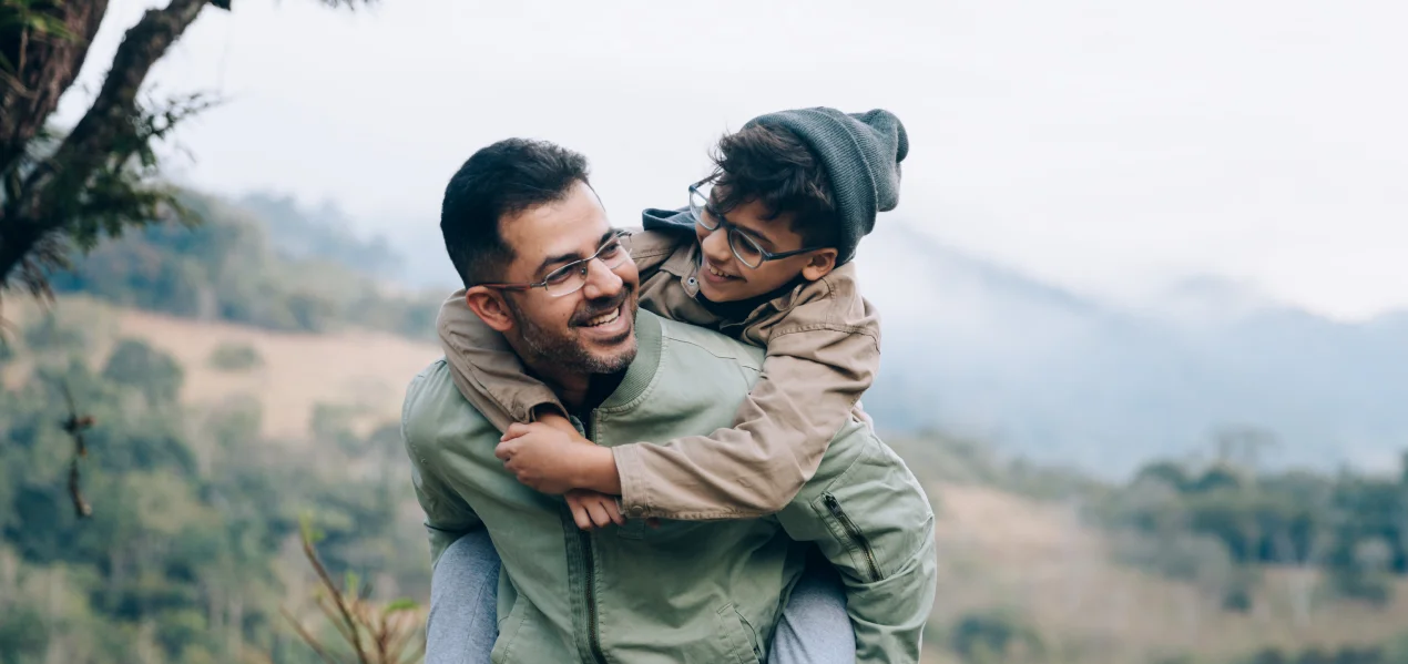A man carrying his son on his back while on a hike on a grassy trail