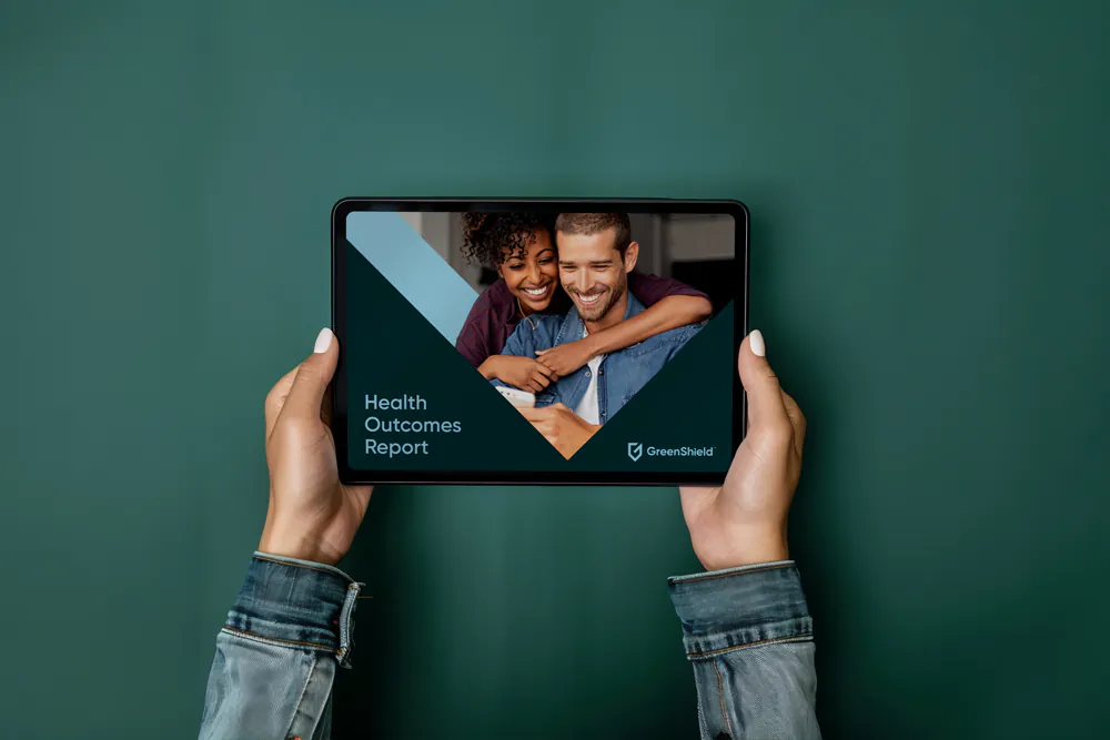 Hands holding a tablet displaying the Health Outcomes Report with a smiling man and woman on the screen, logo of GreenShield at the bottom right.